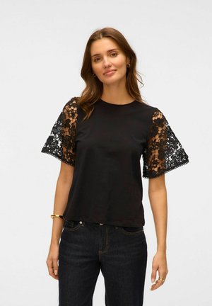 Svart t-shirt med korta spetsärmar som har blommönster, rund halsringning och avslappnad passform, kombinerad med mörka denimjeans.