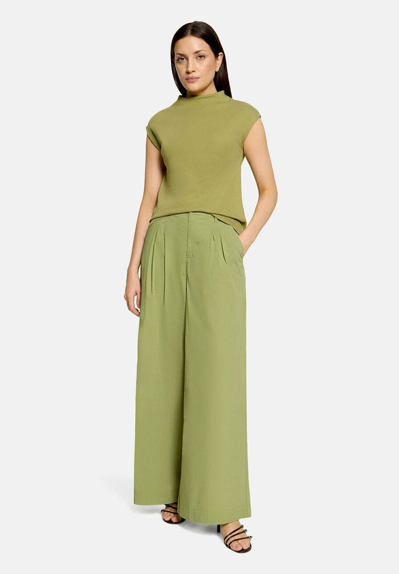 Femme aux cheveux lisses et foncés portant un haut sans manches vert olive, un pantalon large vert clair et des sandales à talons noires sur un fond uni.