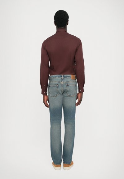 Mörkröd långärmad skjorta, blå denimjeans, bakvy. Jeansen har ljus blekning, kontrasterande sömmar och en brun läderlapp på bakdelen.