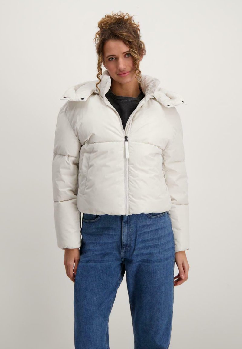 Een vrouw staat tegen een lichte achtergrond, gekleed in een korte witte pufferjack over een zwarte top en blauwe jeans, met een lichte glimlach.