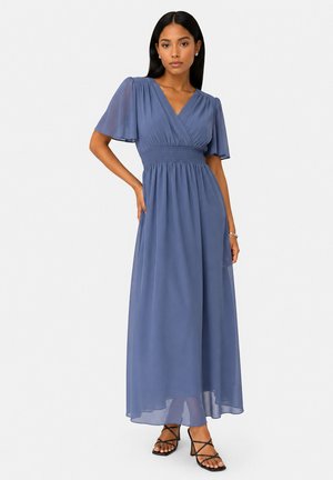 Femme aux longs cheveux foncés portant une robe maxi bleue à col en V avec des manches papillon et des sandales à talons noires à lanières, debout avec une main sur la hanche.
