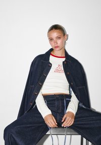 Tummansininen farkkutakki, jossa on erottuva oranssi tikkaus, yhdistettynä valkoiseen pitkähiharaiseen paitaan, jossa on punaisia vivahteita ja printti, sekä yhdistelemä farkkuhousujen kanssa.