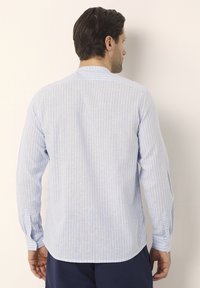 Uomo che indossa una camicia a maniche lunghe di colore azzurro chiaro a righe verticali con colletto alla coreana, visto da dietro contro un muro semplice.