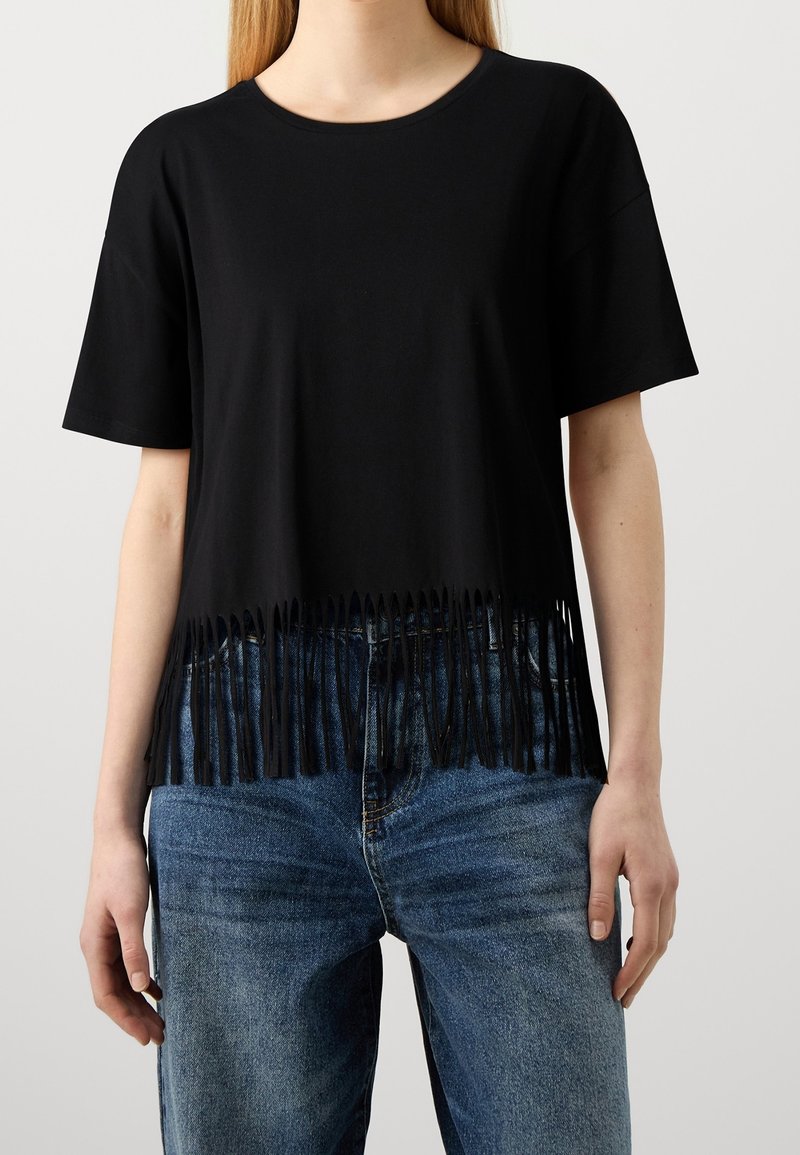 Kvinna som bär en svart t-shirt med kort ärm och fransade nederkant, kombinerad med blå högmidjade jeans mot en enklare bakgrund.