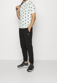 Hombre con zapatillas negras, pantalones jogger negros con un pequeño logo y una camiseta polo de color azul claro con un patrón de perros negros y marrones, sosteniendo una raqueta de tenis.