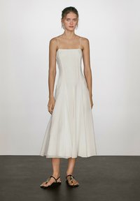 Weißes A-Linien-Kleid mit dünnen Trägern, tailliertem Oberteil und schwingendem Rock. Getragen mit schwarzen flachen Sandalen mit kreisförmigen Akzenten. Glatte Textur.