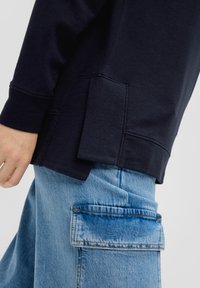 Marineblauwe sweatshirt met een ontspannen pasvorm, voorzien van een zijzak en ribgebreide mouwophangingen. Gecombineerd met lichtblauwe cargobroek, textuur met een patchzak.