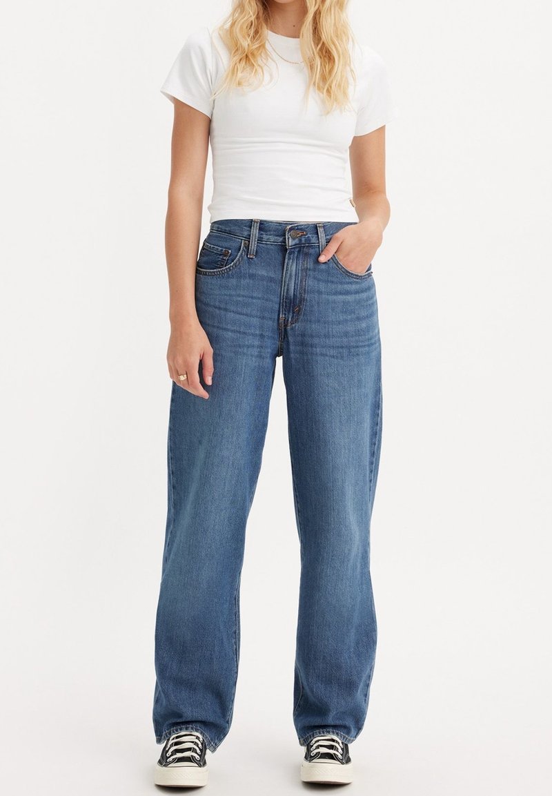 Femme portant une chemise blanche à manches courtes, un jean bleu taille haute à jambes larges, et des baskets noires, debout avec une main dans la poche.