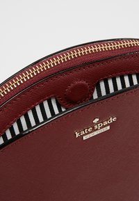 Burgunderfarbene Ledertasche mit goldenem Reißverschluss, ausgestattet mit einem gestreiften Innenfutter. Markenlogo auf der Außenseite eingraviert. Runde Form und elegantes Design.