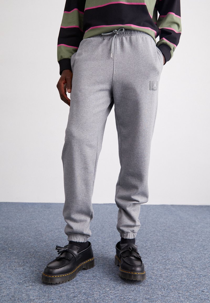 En person står klädd i gråa träningsoverallsbyxor och svarta skor med tofsar, tillsammans med en svart och grön randig sweatshirt. En neutral bakgrund.