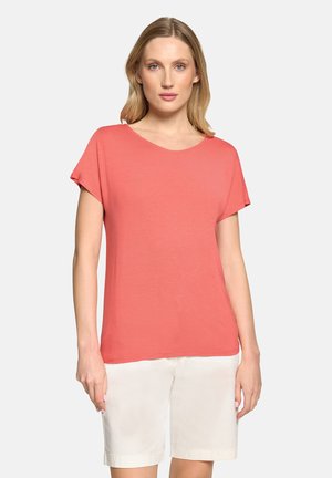 Femme portant un haut corail à manches courtes et un short blanc arrivant aux genoux, se tenant devant un fond clair uni.