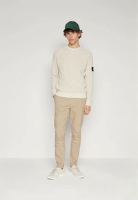 Cream sweater with ribbed texture, tan slim-fit pants, green cap, and white sneakers. Notable logo patch on sweater sleeve.