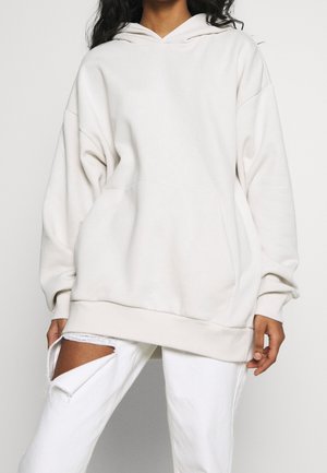 Persona con sudadera con capucha beige de gran tamaño y vaqueros blancos rotos, de pie frente a un fondo claro y liso, con las manos relajadas a los lados.