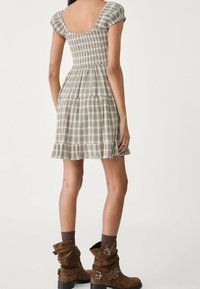 Brązowa krótka sukienka w kratę z prostokątnym tyłem, bufkowymi rękawami i falbaną na dole; w połączeniu z luźnymi brązowymi zamszowymi botkami za kostkę.
