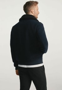 Mężczyzna tyłem, ubrany w ciemnonawigacyjna kurtkę z czarnym kołnierzem z owczej skóry, białą koszulę pod spodem i czarne spodnie, na jednolitym tle.