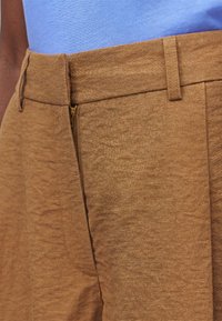 Gros plan d’un pantalon en lin marron avec une braguette ouverte, porté avec une chemise bleu clair rentrée à l’intérieur.