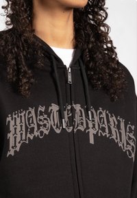 Schwarze Zip-Hoodie mit großem grauen Grafiktext auf der Vorderseite, Kordeln und einer glatten Stofftextur.
