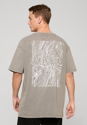 Muž v sivej nadrozmernej tričku s bielym abstraktným vzorom drevených letokruhov a textom „JUST RISE“ na chrbte, otočený bokom.