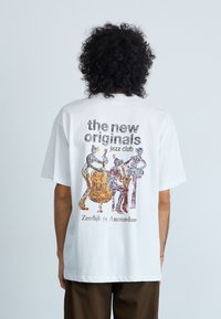 Vit bomull t-shirt med en färgglad jazzklubbsgrafik på baksidan. Texten inkluderar "de nya originalen" och "Zeedijk 59, Amsterdam."