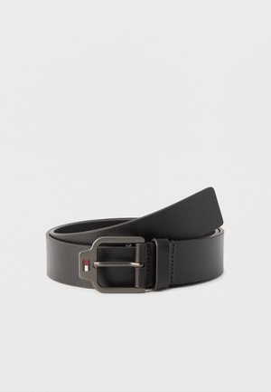 Ceinture en cuir noir avec boucle en métal présentant un petit logo rouge, blanc et bleu, roulée sur un fond blanc.