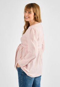 Maglietta premaman texturizzata rosa chiaro con maniche lunghe e cintura annodata, abbinata a jeans blu. Presenta una vestibilità comoda e un design raccolto sulla schiena.