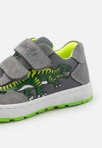 Γκρι αθλητικά παπούτσια για παιδιά με σχέδιο δεινόσαυρου σε πράσινο χρώμα, με επάνω μέρος από δίχτυ, δύο λουράκια με βέλκρο και λευκή σόλα με φωτεινές πράσινες λεπτομέρειες.