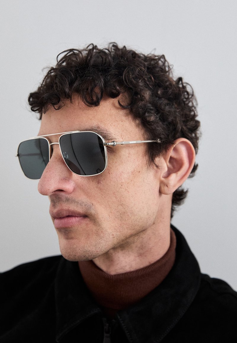 Sunglasses with a silver metal frame, dark lenses, and rectangular shape. The arms feature small cylindrical accents.