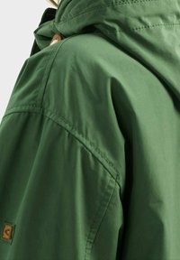 Gros plan d'une personne portant une veste à capuche verte avec des coutures visibles et une petite étiquette de logo en cuir marron.