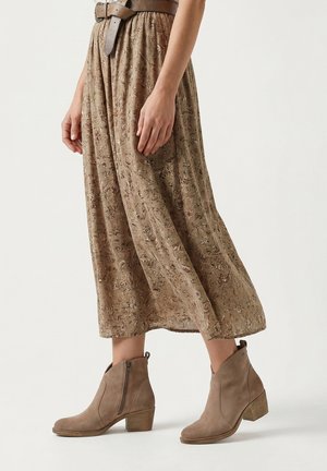 Vrouw draagt een bruine midi-rok met bloemenprint, een brede gestructureerde riem en taupekleurige suède enkellaarsjes met blokhakken tegen een effen achtergrond.