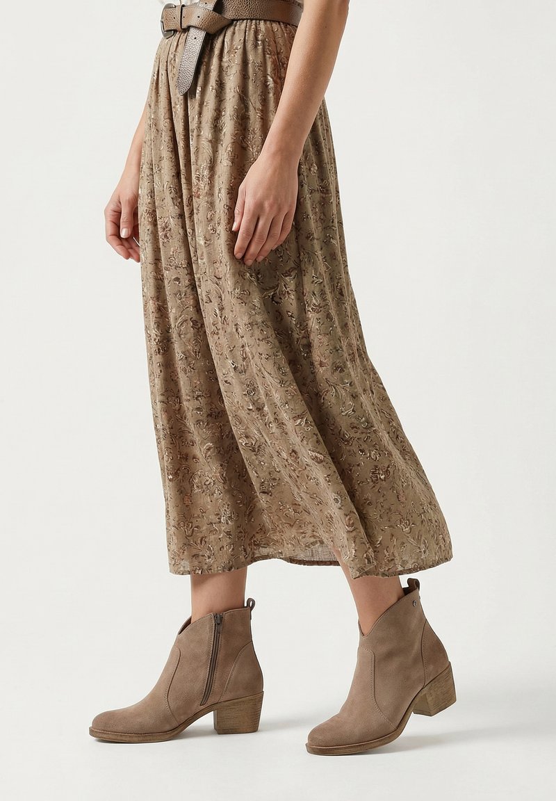 Frau trägt einen braunen, geblümten Midirock, einen breiten, strukturierten Gürtel und taupefarbene Wildleder-Stiefeletten mit Blockabsätzen vor schlichtem Hintergrund.
