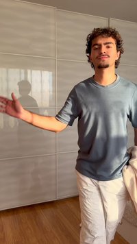 Joven con cabello rizado y perilla, vestido con camiseta gris y pantalones blancos, extiende su brazo en una habitación iluminada por el sol.