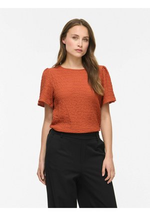 Femme aux longs cheveux bruns portant un haut texturé orange rouille à manches courtes et un pantalon noir taille haute, debout devant un fond blanc.