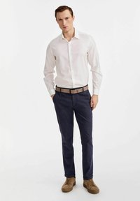 Biała koszula z długim rękawem, ciemnoniebieskie spodnie, brązowy pasek i beżowe buty. Dopasowany krój z kołnierzykiem zapinanym na guziki, teksturowana tkanina i proste linie.