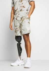 Shorts de camuflaje en beige claro y marrón, combinados con una pierna prostética negra y zapatillas blancas. Diseño casual con bolsillos laterales.