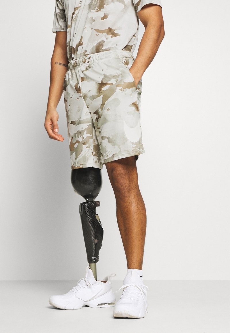 Shorts de camuflaje en beige claro y marrón, combinados con una pierna prostética negra y zapatillas blancas. Diseño casual con bolsillos laterales.