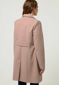 Roze wollen jas met een op maat gemaakte uitstraling, voorzien van een aangeknipte kraag, een rugventilatie en verborgen zijzakken. Gladde textuur, knie-lang.