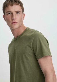 Olivgrünes T-Shirt aus weichem Stoff, mit klassischem Rundhalsausschnitt und kurzen Ärmeln, das eine glatte Textur und einen lockeren Schnitt aufweist.