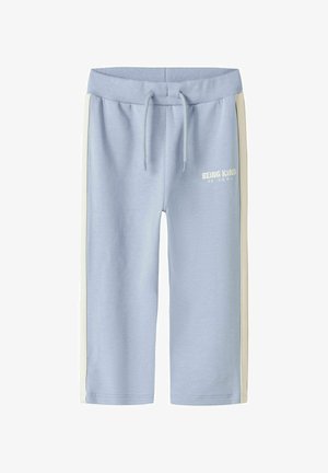 Pantalon de survêtement bleu clair avec des bandes blanches sur les côtés et une taille à cordon, avec le texte "BEING KIND IS COOL" sur la cuisse gauche.