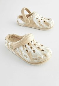 Sabots beiges à motifs avec un intérieur doux en fourrure, des bords surélevés et des trous de ventilation. Équipés d'une sangle à l'arrière pour un maintien sécurisé.