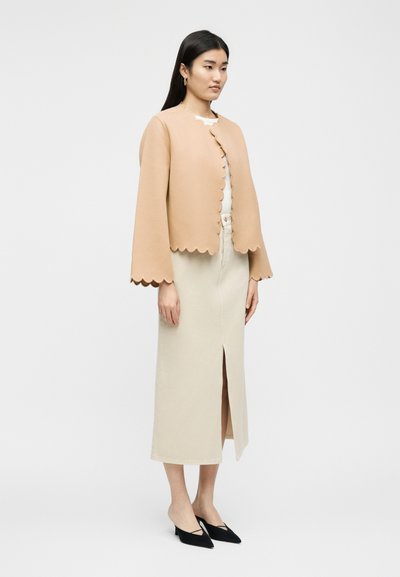 Γυναίκα που φοράει μια μπεζ κοντή ζακέτα με κυματιστά άκρα, ένα λευκό τοπ, μια μίντι κρεμ φούστα με σκίσιμο μπροστά και μαύρα μυτερά τακούνια.