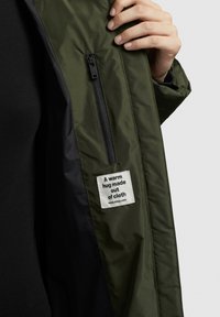 Una mano tiene aperta la manica di una giacca verde, mostrando una tasca con zip interna e un'etichetta bianca che recita "Un caldo abbraccio fatto di tessuto."
