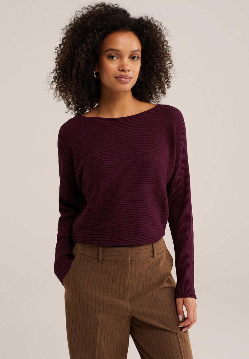 Femme aux cheveux bouclés portant un haut côtelé prune à manches longues et un pantalon marron à rayures fines, mains dans les poches, sur un fond uni.