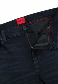 Jeans de mezclilla azul oscuro con una etiqueta roja de HUGO. Presentan un cierre de cremallera, cierre de botón y acentos de hardware metálico. Textura suave.