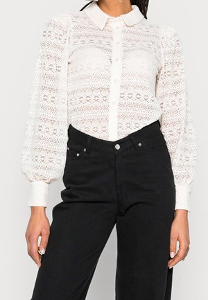 Mujer con una blusa blanca de encaje de manga larga con cuello y pantalones negros de talle alto.