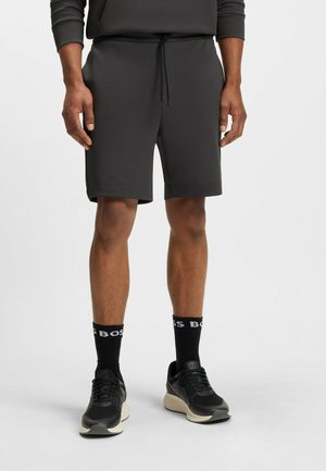 Donkergrijze sportshorts met een trekkoord in de taille, textuurstof en een ontspannen pasvorm, gecombineerd met zwarte sportsokken met een wit logo en zwarte sneakers.