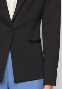 Blazer noir texturé avec un seul bouton sur un top blanc, associé à un pantalon bleu clair, montré du torse jusqu'à mi-cuisse.