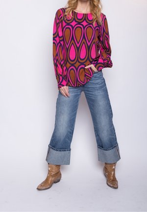 Femme portant un chemisier à manches longues rose et marron à motifs, un jean à jambes larges et revers, ainsi que des bottes de cowboy couleur noisette, debout avec une main dans la poche.