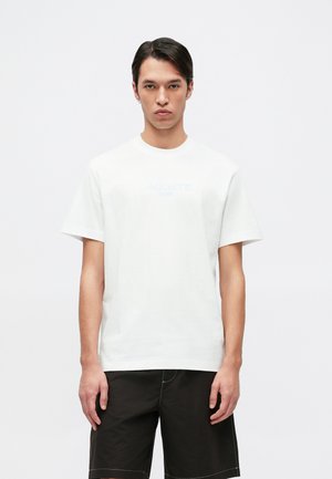 T-shirt bianco in cotone con scollatura rotonda, maniche corte e una stampa blu sottile con la scritta "LACOSTE" al centro. Abbinato a pantaloni corti scuri.