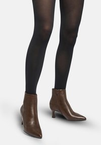 Piernas con medias negras que llevan botas de cuero marrón hasta el tobillo con cremallera lateral y tacones bajos tipo kitten sobre un fondo blanco.