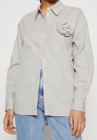 Woman wearing a white button-up shirt with thin black vertical stripes and a fabric flower detail, paired with light blue jeans.
